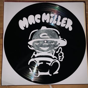 May include: A black vinyl record with the name "MAC MILLER" in white, bold letters. The record features a white cartoon illustration of a character wearing a hat and giving a thumbs-up. The record is in a white sleeve.