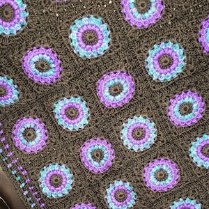 May include: A crocheted blanket with a brown background and purple and blue flowers. The blanket has a border of small blue and purple dots.