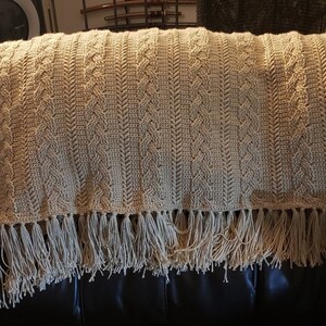 May include: A beige crocheted throw blanket with a fringe border. The blanket has a cable knit pattern and is draped over a brown leather couch.