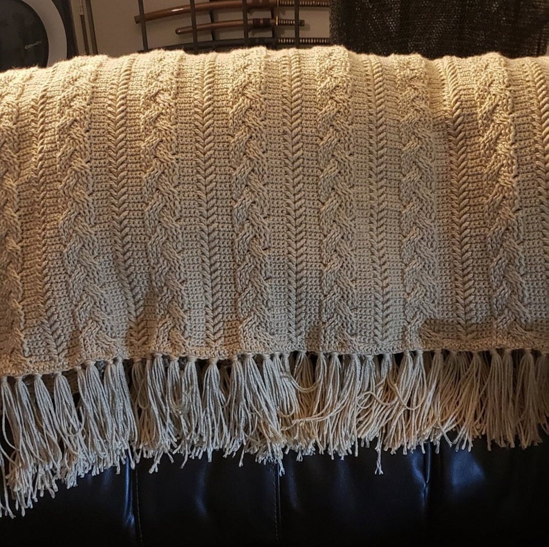 May include: A beige crocheted throw blanket with a fringe border. The blanket has a cable knit pattern and is draped over a brown leather couch.