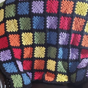 May include: A rainbow granny square crochet blanket with black yarn between each square. The blanket is folded and draped over a black leather chair.