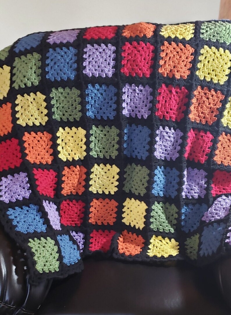 May include: A rainbow granny square crochet blanket with black yarn between each square. The blanket is folded and draped over a black leather chair.