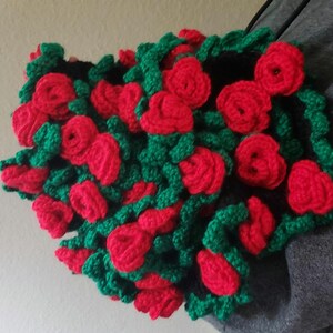 May include: A handmade crochet floral accessory featuring vibrant red roses and green leaves. The roses are densely packed, creating a textured, three-dimensional effect. The piece is likely a decorative item or wearable accessory.