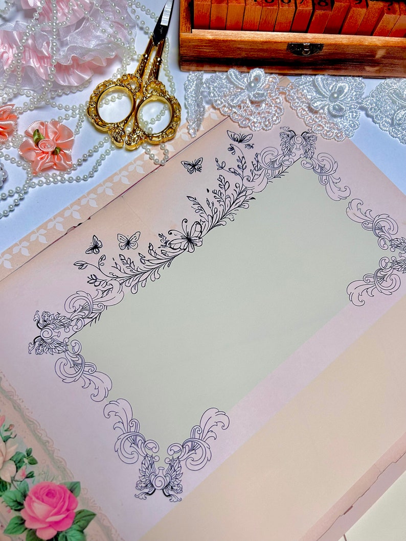 K&ouml;nnte beinhalten: Eine leere Seite mit einem dekorativen Rand aus schwarzen floralen Designs und Schmetterlingen. Die Seite ist umgeben von wei&szlig;er Spitze, Perlen und rosa Stoffblumen.