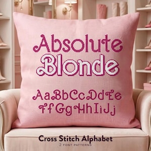 May include: A square pink throw pillow featuring dark pink cross-stitch embroidery. The text reads Absolute Blonde in a stylised font, followed by the alphabet letters Aa through Jj in the same dark pink cross-stitch pattern.
