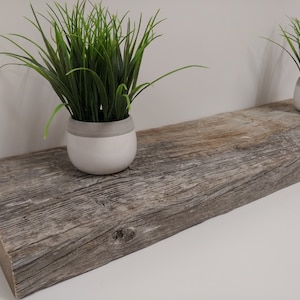 May include: A rustic wooden shelf with a weathered gray finish. Two small white pots with a gray band and green grass plants sit on the shelf. The shelf is made of a thick, rough-hewn plank.