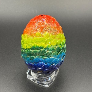 May include: An egg-shaped object covered in overlapping, scale-like textures in a vibrant rainbow gradient of red, orange, yellow, green, blue, and purple. The surface is coated in a fine, shimmering gold glitter finish and rests on a clear stand.