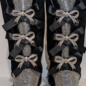 May include: Pair of black boots with rhinestone accents and black satin bows. The boots have a faux fur trim at the top and rhinestone detailing on the front and back. The bows are embellished with rhinestones.