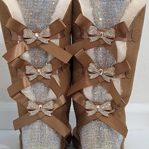 May include: A pair of brown suede boots with a sparkling rhinestone panel on the back and a white faux fur trim. The boots are adorned with brown satin bows and rhinestone bow accents.