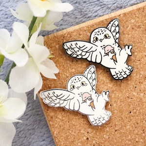 May include: Two white enamel pins of a cartoon snowy owl holding an ice cream cone. The owls have black eyes and beaks, and the ice cream is pink and white.