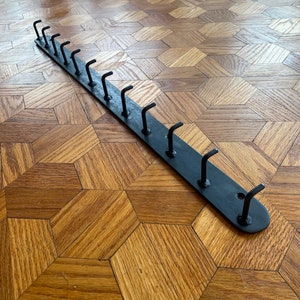 May include: A black metal hook rack with 10 hooks. The rack is about 18 inches long and is lying on a wooden floor.