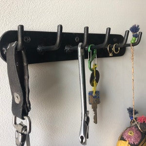 May include: Black metal wall mounted hook rack with six hooks. A leather strap with a metal clip, a wrench, a carabiner, a key ring with a key and a small round tag, and a string with a small colorful fabric ball are hanging from the hooks.