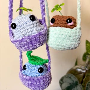 May include: Three handmade crochet plant hangers, each with a unique character. One is light blue, one is brown, and one is blue. Each has large black eyes and a small green plant sprouting from the top. The hangers are purple, mint green, and light green.