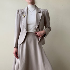 May include: A beige skirt suit with a fitted blazer and a slightly flared skirt. The blazer has a single button closure and a gold pin on the lapel.