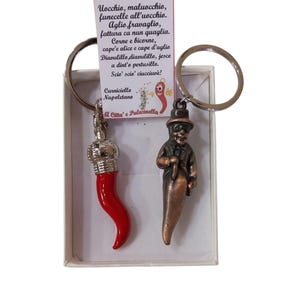 Könnte beinhalten: Zwei Schlüsselanhänger mit Anhängern. Einer ist eine rote Chilischote mit einer silbernen Krone, der andere ist eine kleine Figur mit Hut und Chilischoten-Körper. Eine Karte mit italienischem Text befindet sich im Hintergrund.