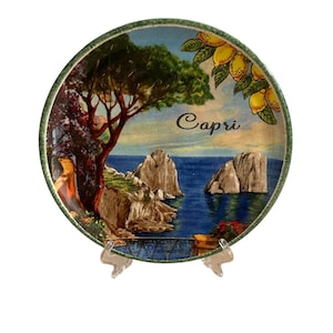 Può includere: Un piatto rotondo in ceramica con una scena dipinta dell'isola di Capri, Italia. Il piatto presenta un bordo verde, un mare blu, un albero verde e due isole rocciose. La parola "Capri" è scritta in verde sul piatto.