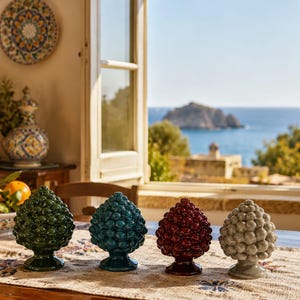 Peut inclure: Quatre sculptures en céramique en forme de pomme de pin, vertes, turquoise, bordeaux et grises, sont posées sur une table. Les sculptures ont une surface texturée et reposent sur un socle. L'arrière-plan montre une fenêtre avec vue sur l'océan et une petite île.