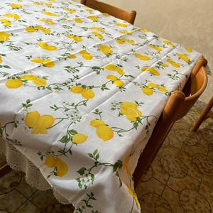 May include: White tablecloth with a lemon print. The tablecloth has a lace trim and is on a wooden table with brown chairs.