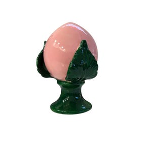 Könnte beinhalten: Eine Keramik-Pfirsich-Skulptur mit glänzendem rosa Finish. Der Pfirsich wird von dunkelgrünen Blättern umschlossen und sitzt auf einem grünen Sockel. Die Skulptur ist ein dekoratives Objekt.