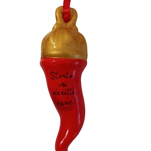 Può includere: Un ciondolo a forma di corno italiano in ceramica rossa con la parte superiore dorata e un nastro rosso per appenderlo. Il ciondolo riporta la scritta "Storta va diritta vene!" in inchiostro nero.
