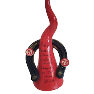 Può includere: Un corno di toro in ceramica rossa con anelli a forma di ferro di cavallo neri e dorati. Gli anelli hanno i numeri "5" e "73" su di essi. Il corno ha il testo "Fortuna, Successo e olemnto a volonta chistu l'auguro pe sta muova attivita!" scritto su di esso.