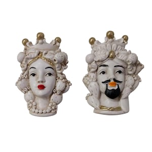 Può includere: Due sculture di teste in ceramica, una femminile e una maschile, con dettagli bianchi e dorati. La testa femminile ha labbra rosse, una corona e una collana floreale. La testa maschile ha barba e baffi neri e bocca arancione. Entrambi hanno i capelli ricci.