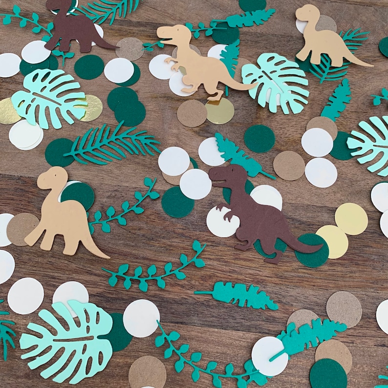 Op de afbeelding: Groene, bruine en witte confetti met dinosaurussen en bladeren. De confetti ligt op een bruin houten oppervlak.