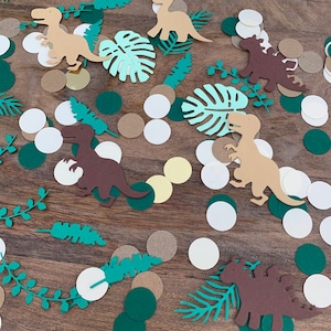 Op de afbeelding: Een verzameling groene, bruine en witte papieren uitsnijdingen van dinosaurussen, bladeren en cirkels. De uitsnijdingen zijn verspreid over een bruin houten oppervlak.
