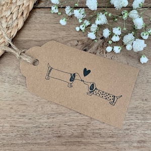 Op de afbeelding: Een bruin papieren cadeau-etiket met twee zwart-witte teckels die elkaar kussen. De honden dragen truien met hartjes erop. Boven de honden is een zwart hartje.