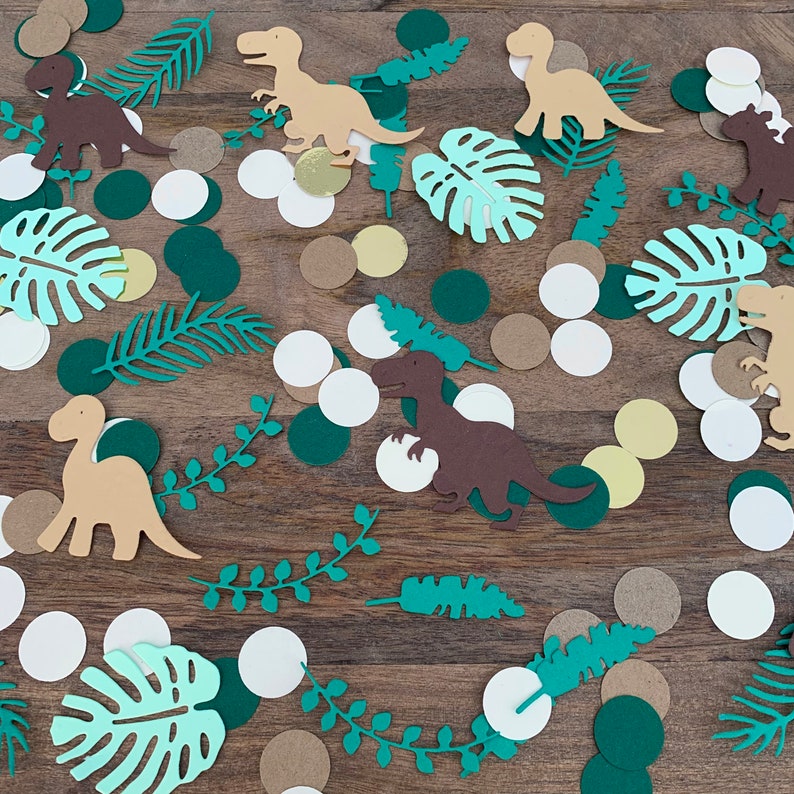 Op de afbeelding: Een verzameling groene, bruine en gouden confetti met dinosaurussen en tropische bladeren. De confetti is perfect voor een dinosaurus-themafeest.