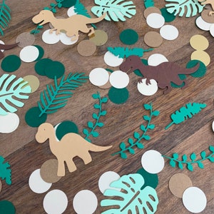 Op de afbeelding: Groene, bruine en witte confetti met dinosaurussen en bladeren. De confetti ligt op een bruin houten oppervlak.