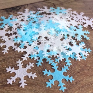 Op de afbeelding: Een stapel witte en lichtblauwe vilt sneeuwvlokken uitsnijdingen. De sneeuwvlokken zijn verspreid over een houten oppervlak.