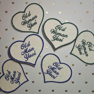 May include: A collection of heart-shaped patches. Some patches have a white background with blue trim and the text "Old Man Yaoi" embroidered in blue. Other patches have a white background with green trim and the text "Old Woman Yuri" embroidered in green.