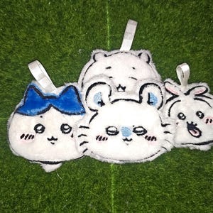 May include: A set of four white plush ornaments with embroidered facial features and small fabric hanging loops. The designs include a character with a blue bow, a cat-like figure, a bear-like face, and a small round creature with pink ears.