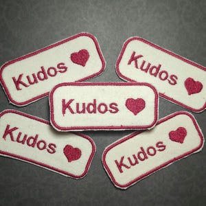Puede incluir: Cinco parches bordados con la palabra "Kudos" en escritura burdeos, cada uno con un corazón burdeos. Los parches son rectangulares con esquinas redondeadas, bordeados en burdeos, sobre un fondo crema.