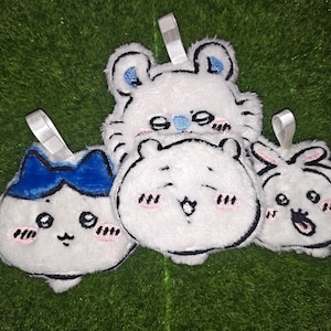 May include: Four white plush ornaments with embroidered facial expressions and small white fabric loops for hanging. The designs include a character with a blue bow, a bear-like figure, a round face with a wide smile, and a surprised expression.