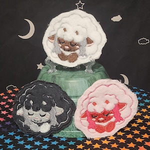 Puede incluir: Tres parches bordados con forma de oveja en blanco, negro y rosa. Cada parche presenta una cara de oveja con los ojos cerrados y un contorno esponjoso en forma de nube. Los parches se exhiben sobre un pedestal verde sobre un fondo estrellado.
