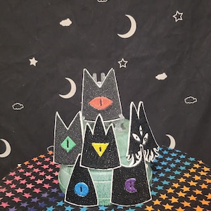 Pode incluir: Cinco patches de feltro preto em forma de gato com olhos de diferentes cores. Os olhos são vermelhos, verdes, amarelos, azuis e roxos. Os patches estão sobre um fundo de estrelas brancas sobre uma superfície preta.