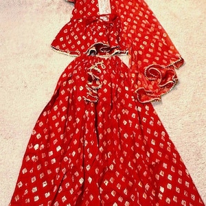 May include: A red outfit comprising a top, skirt, and shawl. The fabric features a pattern of small, silver diamond shapes. The skirt and top edges are trimmed with gold-coloured detailing.