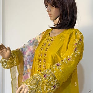 Könnte beinhalten: Senfgelbe bestickte Tunika mit floralen Details an den Ärmeln und am Oberteil. Die Tunika hat eine aufwendige weiße und kastanienbraune Blumenstickerei. Ein durchsichtiger Blumenschal ist über die Schultern drapiert.