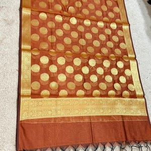 May include: A rust-colored textile with a gold floral pattern and a gold border. The fabric has a sheer quality, and the bottom edge is finished with black tassels. The design features repeating gold circular floral motifs.