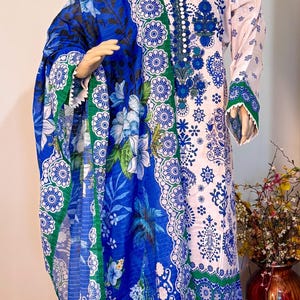 Peut inclure: Un salwar kameez rose clair et blanc à imprimé floral avec un dupatta floral bleu et vert assorti. La tenue présente des motifs floraux bleus complexes et une bordure verte. L'ensemble est présenté sur un mannequin.