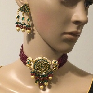 May include: A gold choker necklace with a round pendant featuring green and white stones. The pendant is surrounded by red beads and has a dangling chain of green, red, and white beads. The necklace is worn by a mannequin.