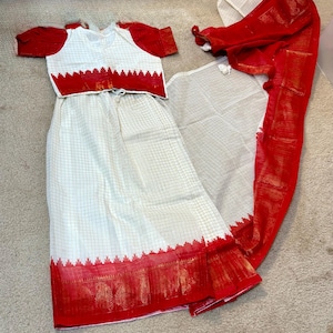 May include: A traditional Indian outfit featuring a white skirt and top with a red border and accents. The top has short sleeves and a matching red border. A red and white scarf is also included.
