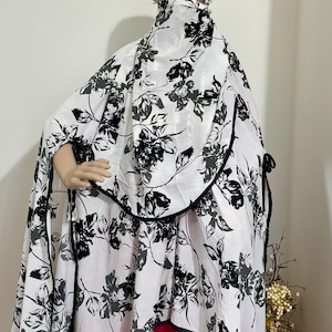 May include: A white and black floral patterned garment, including a head covering and cape, is displayed on a mannequin. The cape is trimmed with black piping and ties. A bright pink dress is visible underneath.