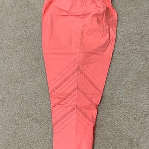 May include: A pair of coral pink pants with a decorative lace trim on the bottom. The pants have a wide waistband and a straight leg.