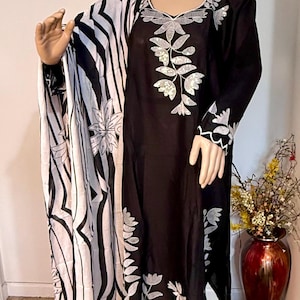 May include: Black dress with white floral embroidery and a matching black and white patterned shawl. The dress features long sleeves and a square neckline. The mannequin is standing on a dark gray platform.