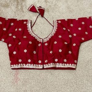 May include: A maroon blouse with short sleeves and a deep V-neck. The blouse is adorned with silver embroidery along the edges and scattered floral patterns. The fabric has a slight sheen.