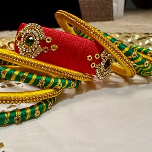 以下が含まれることがあります： カラフルなバングルとカフブレスレットのコレクション。バングルは黄色と緑で、ゴールドのアクセントが付いています。カフブレスレットは赤色で、ラインストーンと中央に緑色の石が飾られています。ジュエリーは金色のトレイに展示されています。