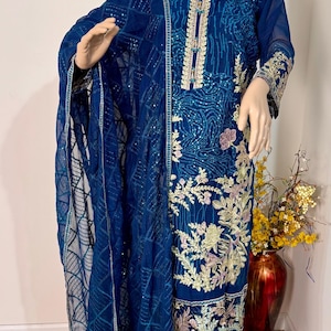 May include: A blue and gold embroidered dress with a matching dupatta. The dress has a long-sleeved design and a floral pattern. The dupatta is sheer and has a geometric pattern.